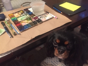 A small Cavalier King Charles Spaniel dog sits on a chair in front of a watercolor workstation
