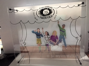 A sculpture shows a painting of four young people gesturing toward Ames Children's Theater logo with theater curtains represented in a foreground layer
