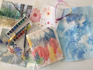 Watercolor paints and sketches arrayed on a tabletop with a pair of colorful glasses