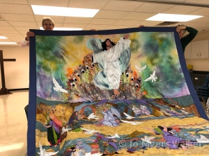 Skilled assistants hold up revised Easter story wall hanging by artist Jo Myers-Walker showing the emphasized figure of Jesus