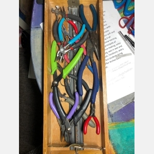 An assortment of many different pairs of pliers, many with colorful handles, jumbled in a narrow wooden drawer from a card catalog