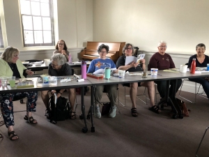 A class of artists seated at a long table all facing the camera. Bright sunlight is coming through the window behind them