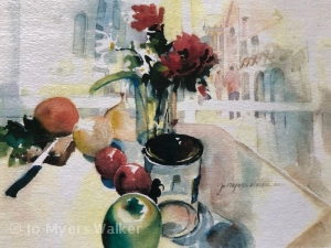 Watercolor still life by artist Jo Myers-Walker of a tabletop featuring a small vase with red flowers and daisies and produce including onions, an apple, and greens next to a knife and empty jar. Buildings with archways are faintly visible in the background.