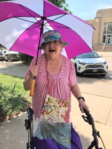 Artist Jo Myers-Walker in sunny downtown Iowa City holding a pink and white striped parasol and wearing patchwork sun hat and purse which she sewed